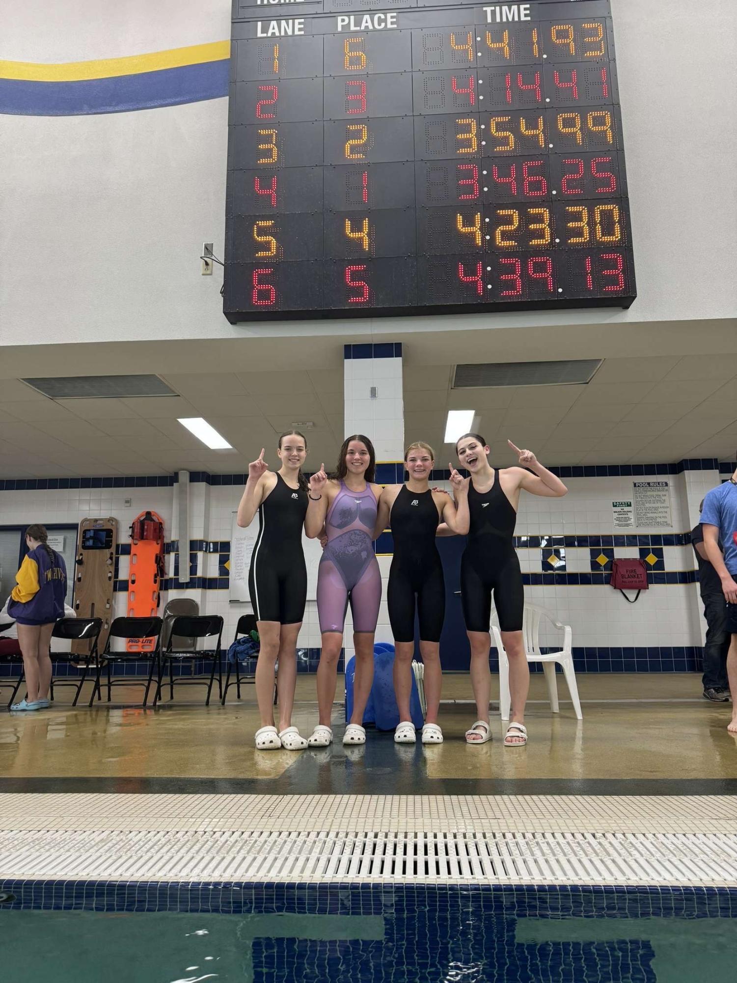 Left to right: Anna Schliepp, Alexia Pacurar, Payton Zamzow, and Caliber Hartzke stand in front of the scoreboard after taking first in their heat in the 400 freestyle relay