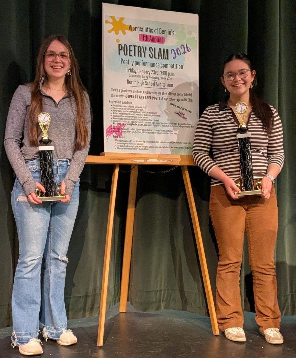  Runner-Up Ella Spitler (left) and First place winner Tasi Ana Sanchez (right) stand next to the Poetry Slam poster after the competition. “I submitted two poems, one about a lost flower, and another about rain,” Spitler said. 
